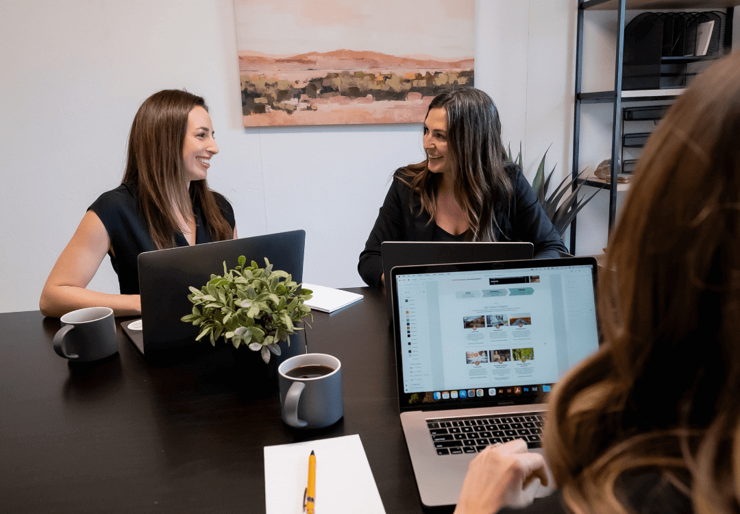 202207_OTM_BlogGraphics Three women in a business meeting, two smiling at each other, around a table with laptops, a notepad, pens, and coffee mugs while discussing hiring a marketing agency. A landscape painting hangs