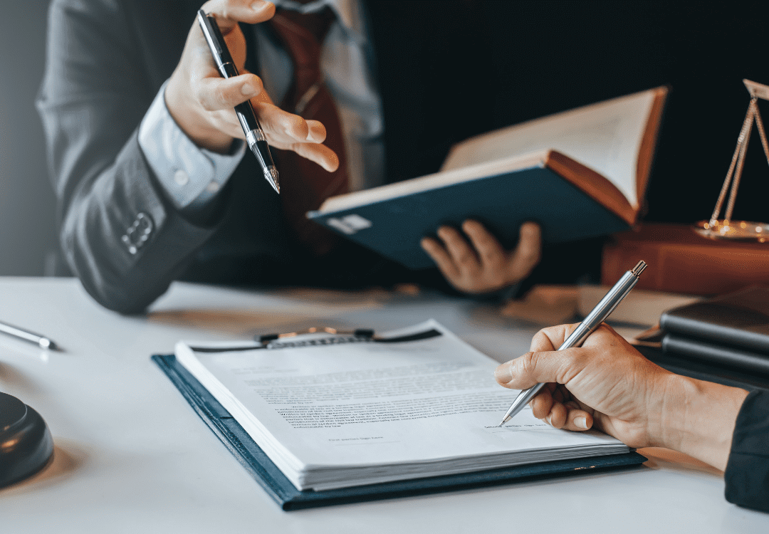 close-up-of-legal-team-working-on-paperwork Two people are engaged in a discussion or meeting. One is holding an open book and gesturing with a pen, while the other is signing a document on a clipboard. A gavel and scales of justice are visible on the desk, indicating a legal or professional setting.