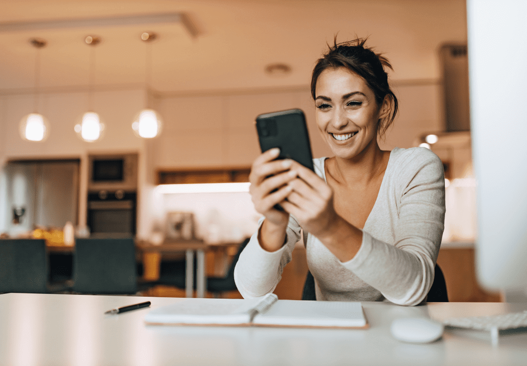 young-woman-looking-at-phone-and-smiling A woman with a smile is holding her smartphone while sitting at a desk. She is in a modern kitchen with pendant lights hanging from the ceiling, and there is a notebook and pen on the desk in front of her.
